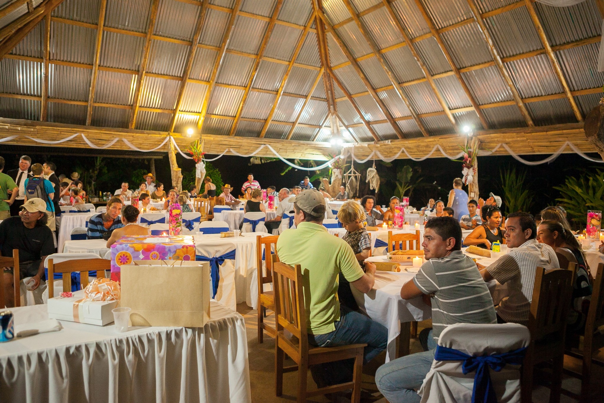 Community gathering at a large, open-air event under a thatched roof.