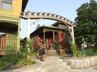 Person standing in front of a small house with a wooden archway and garden.