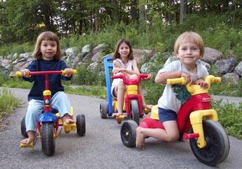 Three children riding tricycles outdoors on a paved path.