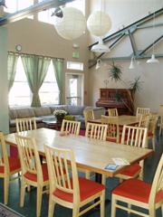 Bright communal dining area with wooden tables and chairs, green curtains, and hanging lights.
