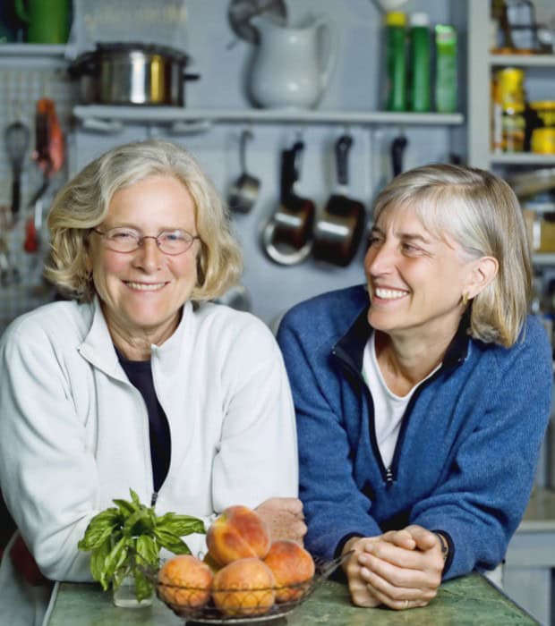 Two women smiling in a kitchen with fresh peaches and herbs on the table.