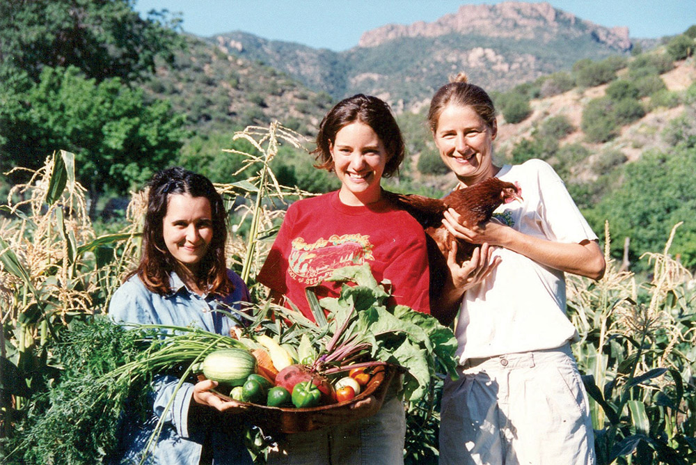 Three women in a farm with vegetables and a chicken, mountains in the background.