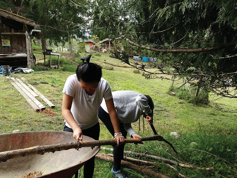 People working outdoors in a rural setting with trees and wooden structures.