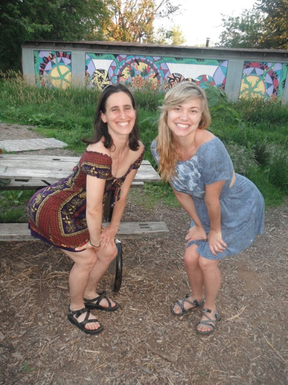 Two women smiling outdoors in front of a colorful mural at an intentional community.