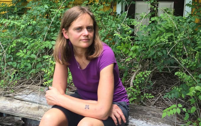 Woman sitting outdoors on a wooden bench surrounded by green foliage.