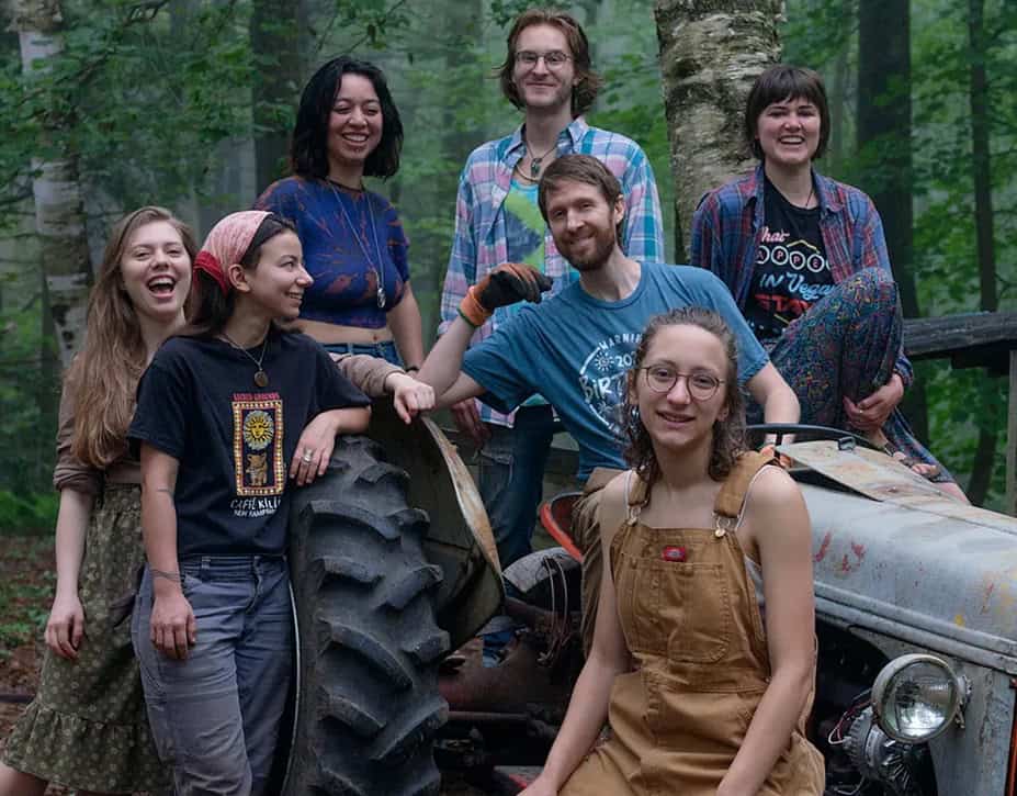 Group of diverse young adults smiling outdoors with tractor in a forest setting.