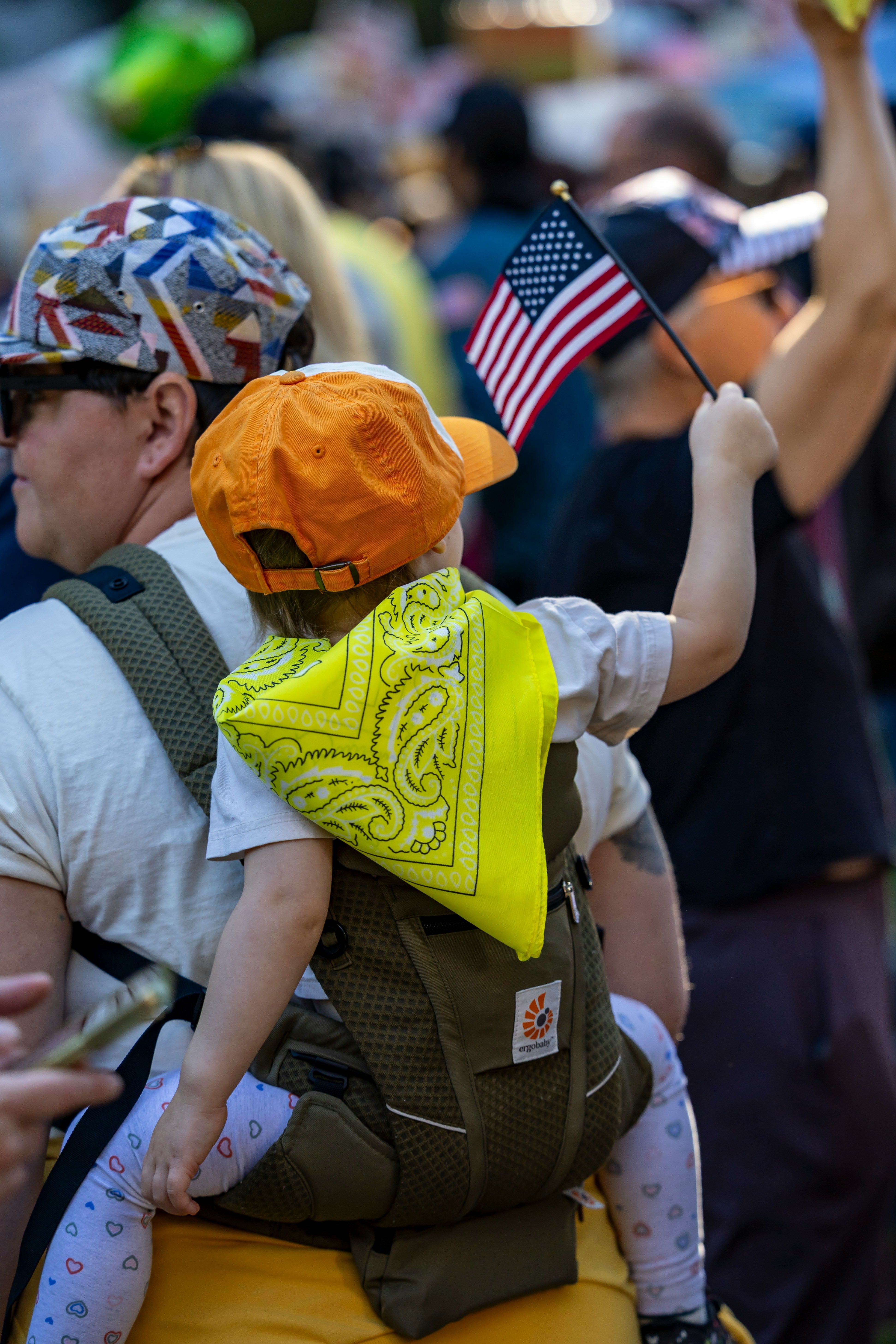 Child with orange hat and yellow bandana holding American flag at community gathering.