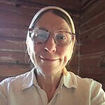 Woman with glasses smiling in a wooden cabin, casual attire.