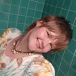 Close-up of a smiling woman with short hair and floral blouse in front of green tiled wall.