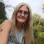 Portrait of a woman with long blonde hair and glasses outdoors near water.