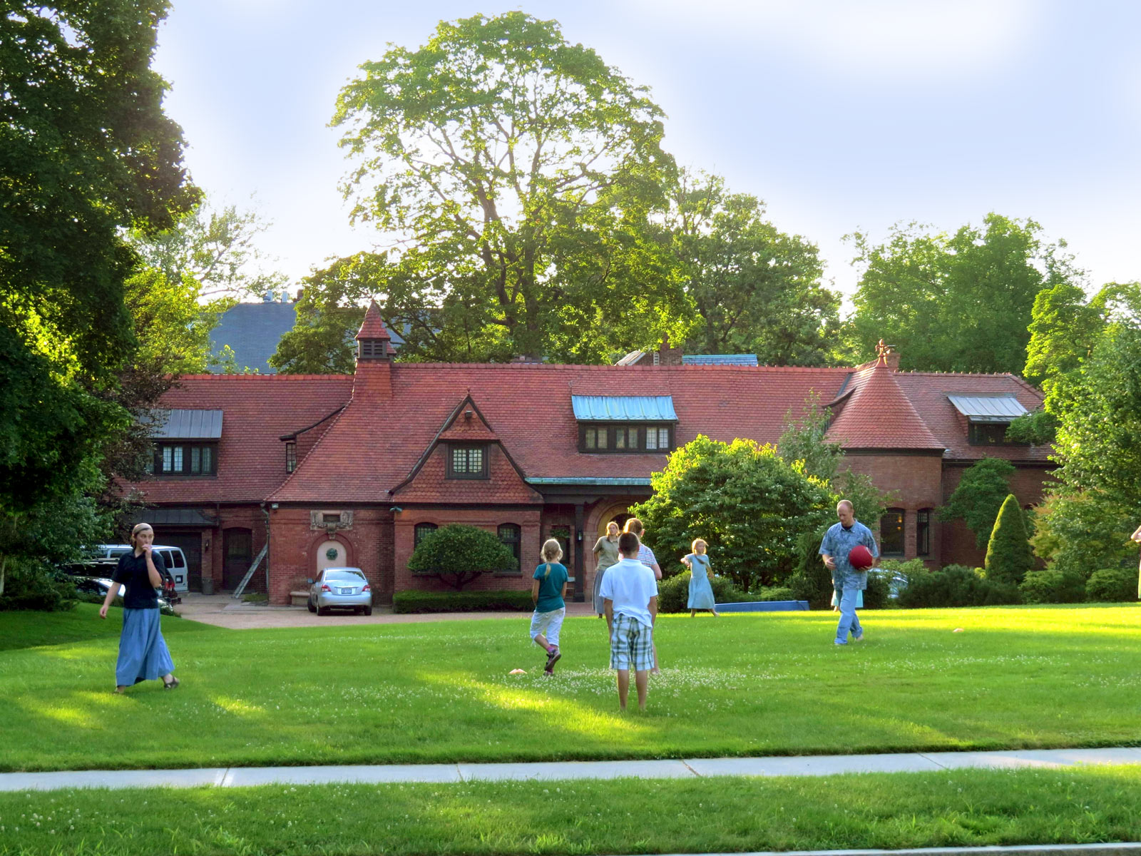 People walking on grass near a historic community building with trees.