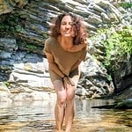 Woman smiling at a waterfall in a natural setting with rocks and greenery.
