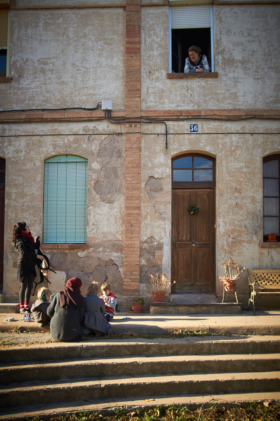 People sitting outside a rustic house with a woman looking out from the window.