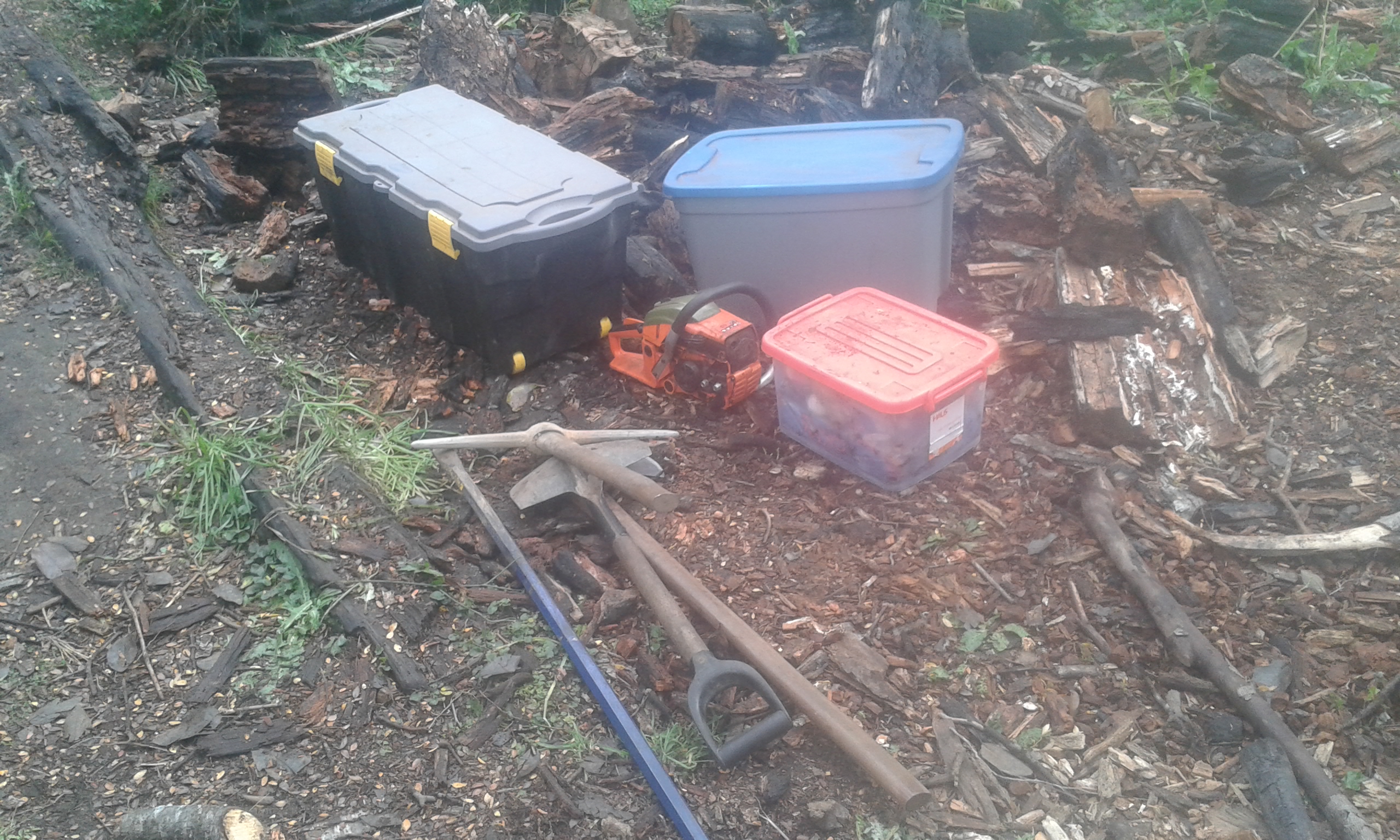 Tools and storage containers placed on forest ground with soil and wood debris.