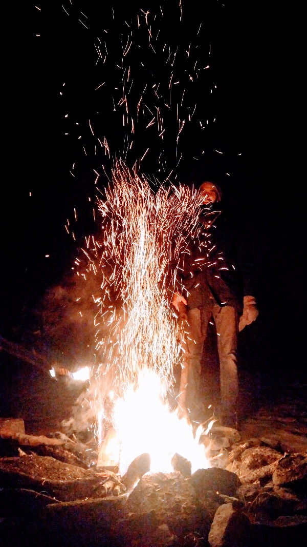 Person standing near a campfire with sparks flying into the night sky.