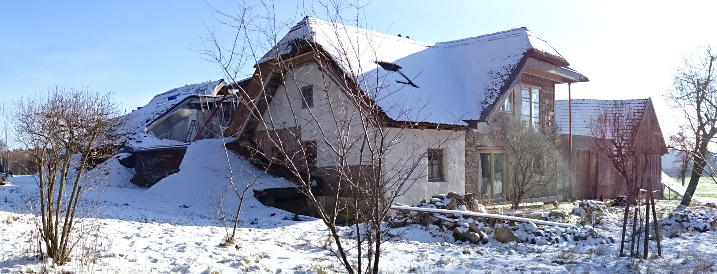 Snow-covered house with trees and clear blue sky in a rural area.