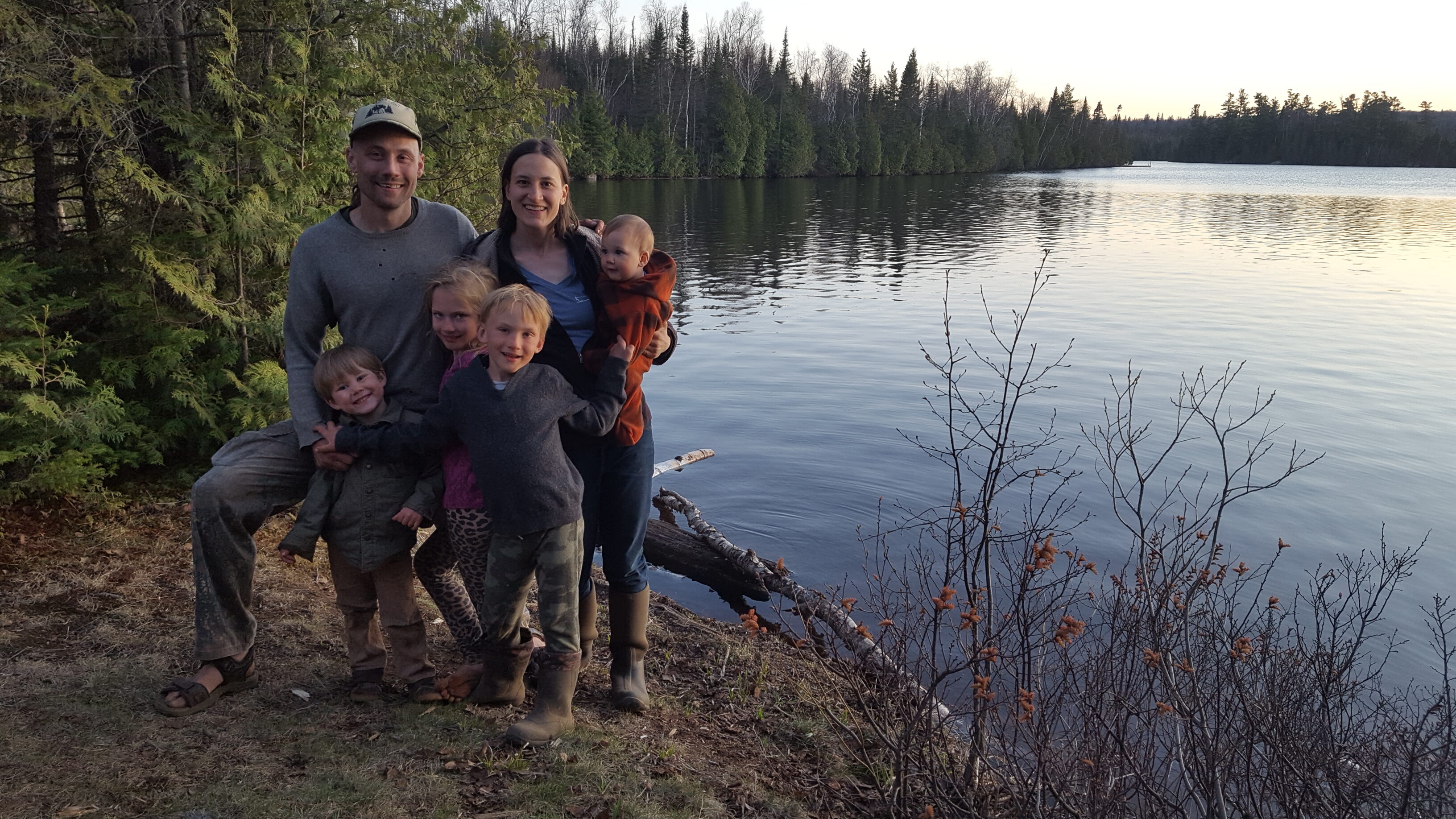 Family of six by a lake with trees and calm water in the background.