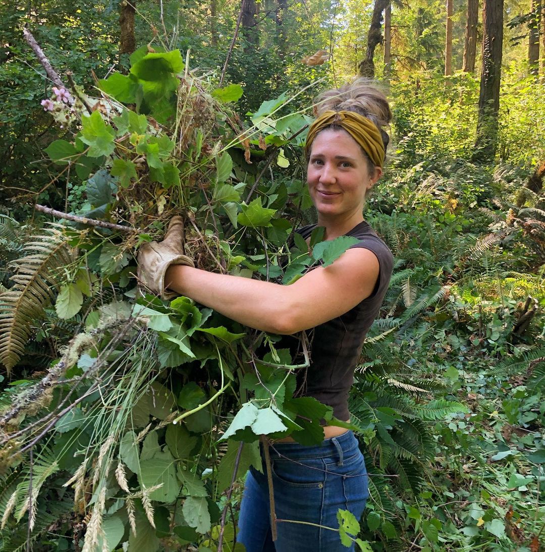Woman holding a large plant in a lush forest setting.