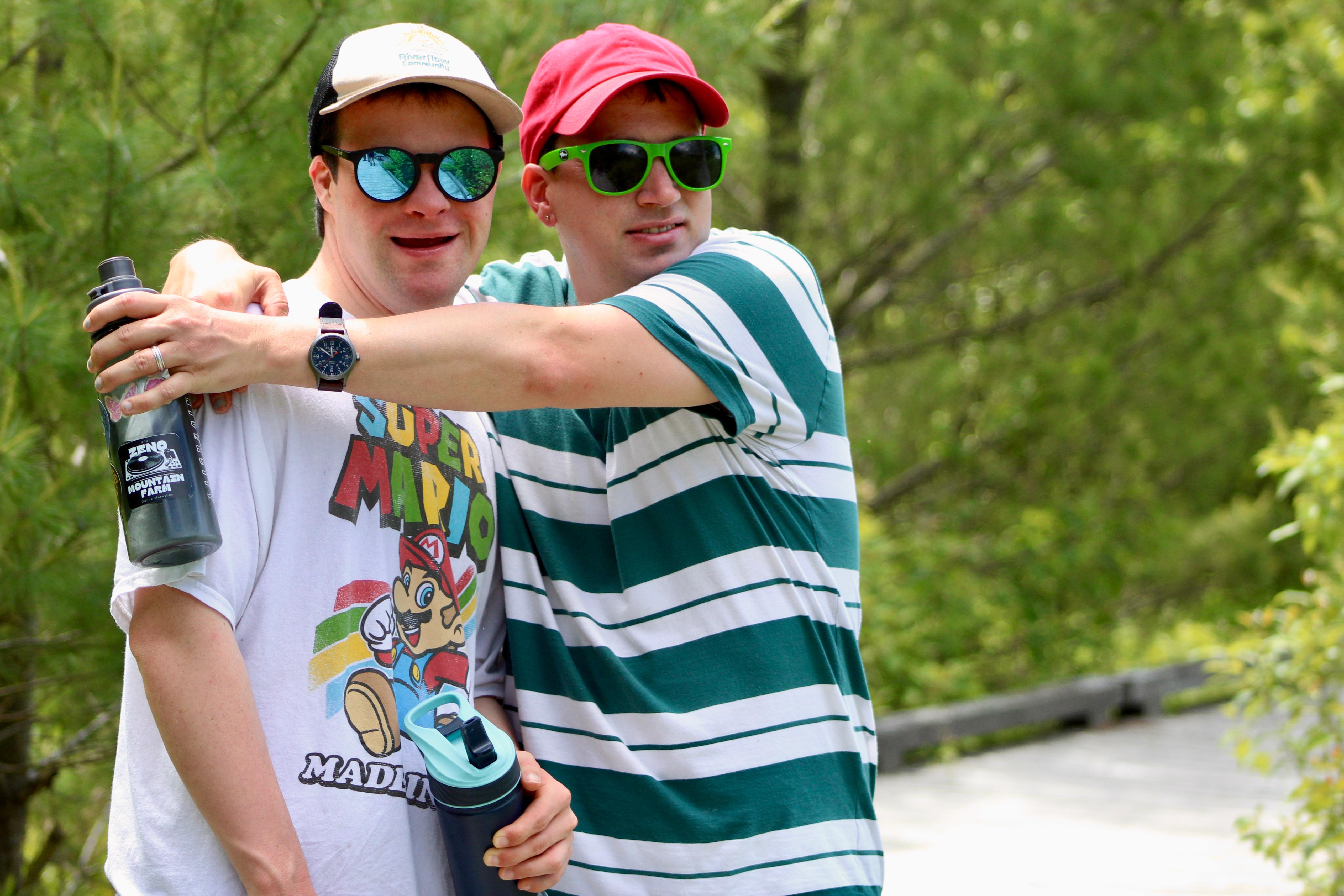 Two young men in casual clothing and sunglasses embracing outdoors in a green setting.