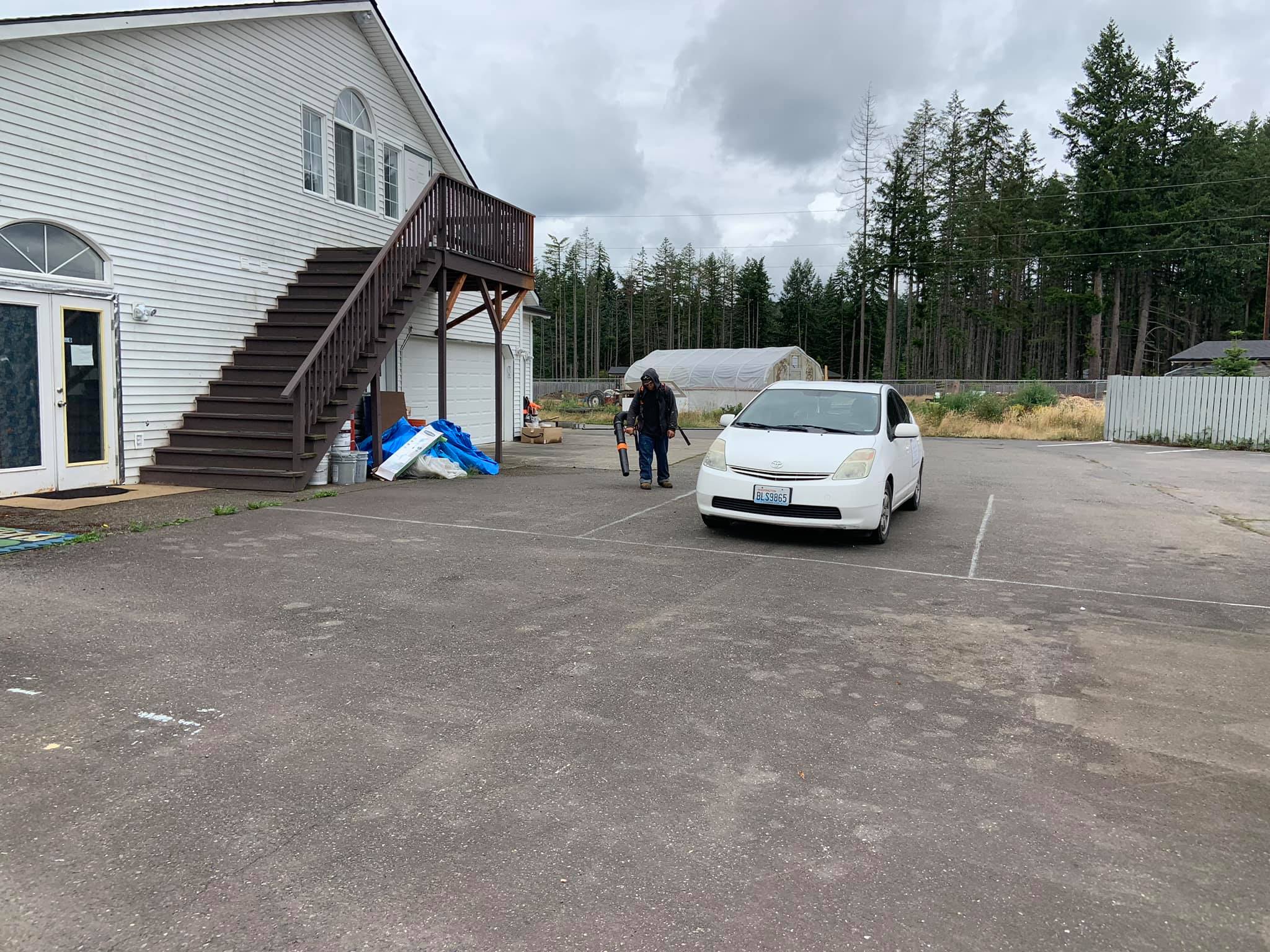 Parking lot with white car and two people near greenhouse and house.