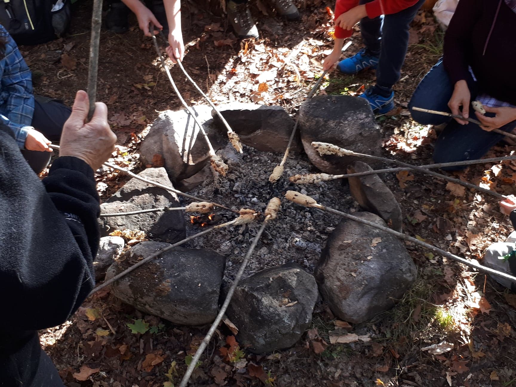 People gathered around a campfire cooking with sticks in a community setting.