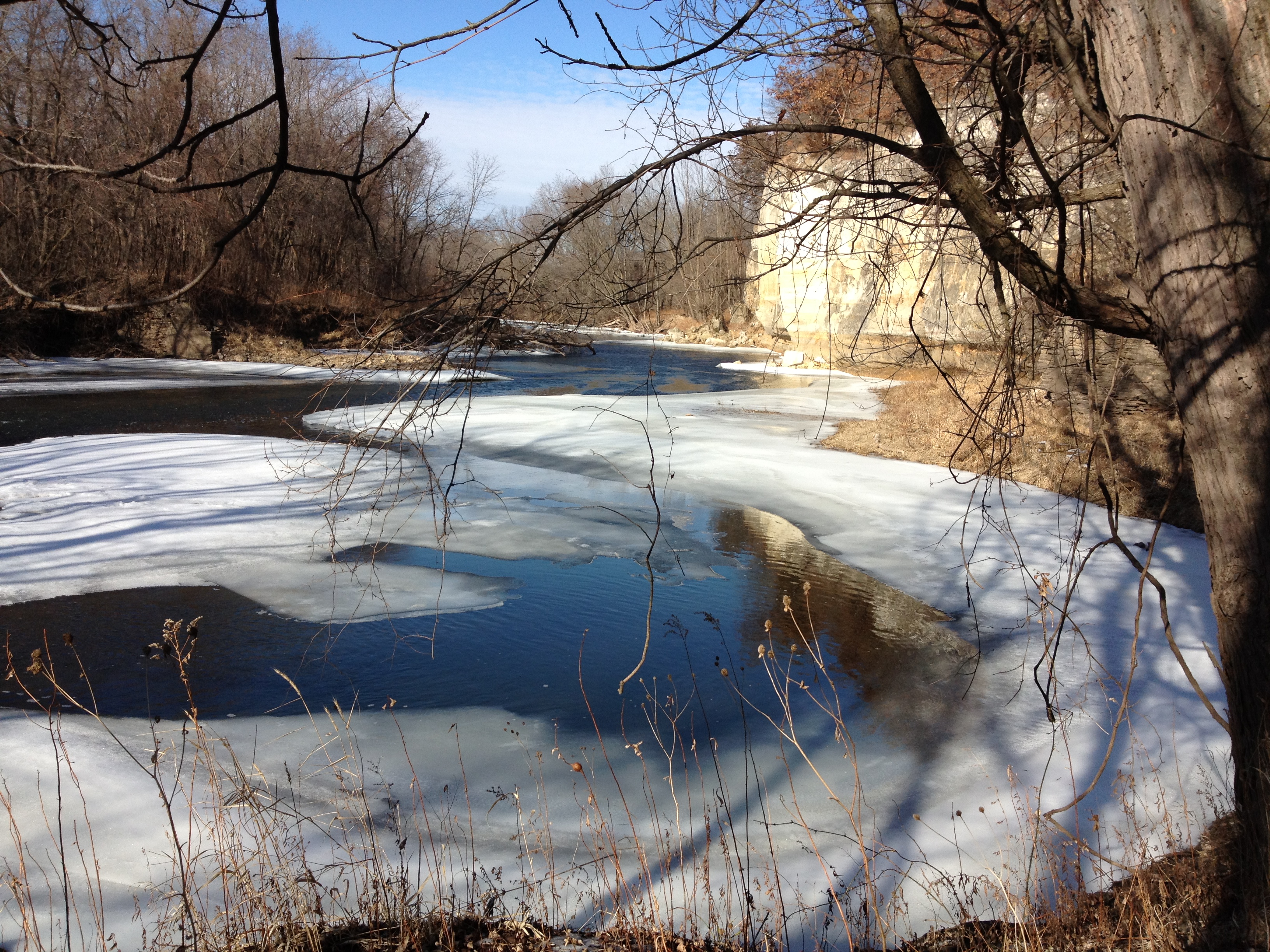 Frozen river with ice and snow, surrounded by leafless trees and cliffs.