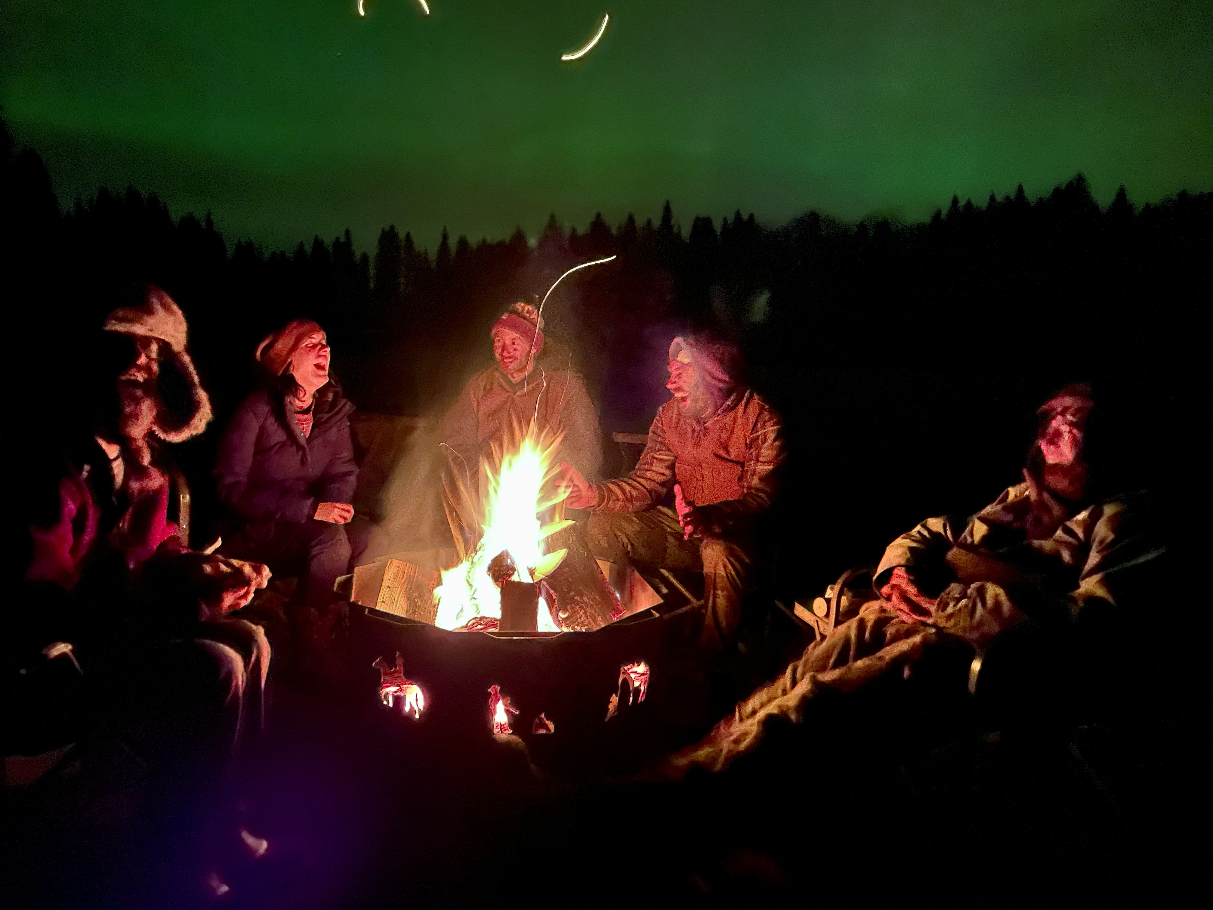 People sitting around a campfire under the Northern Lights at night.