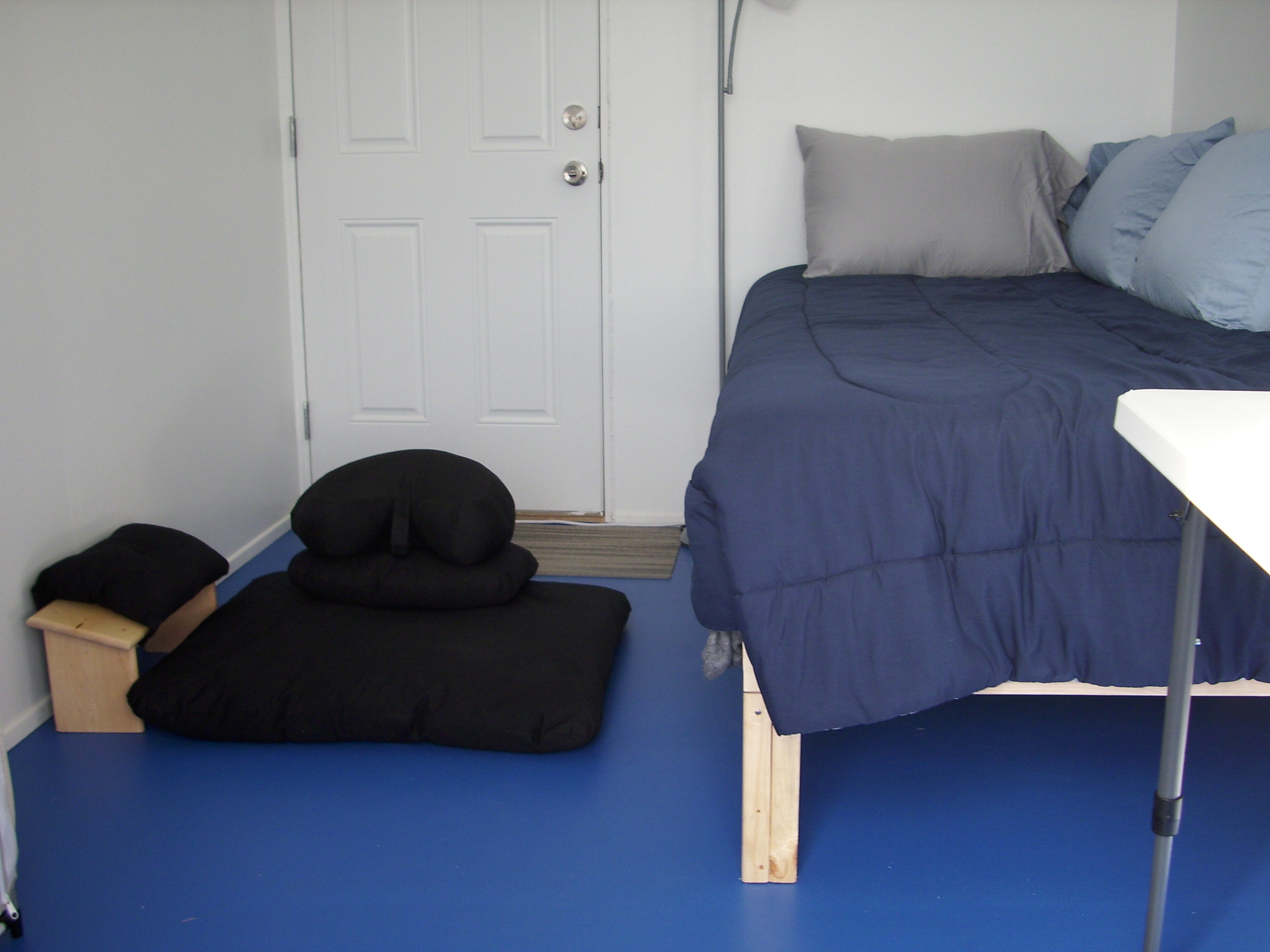 Minimalist bedroom with bed and meditation cushion on blue floor.
