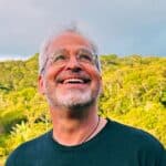 Smiling man outdoors with lush green background and cloudy sky.