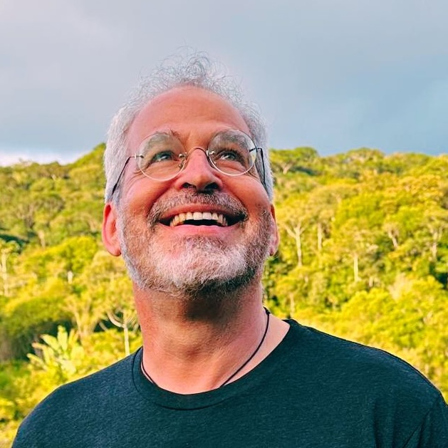 Smiling man outdoors with lush green background and cloudy sky.