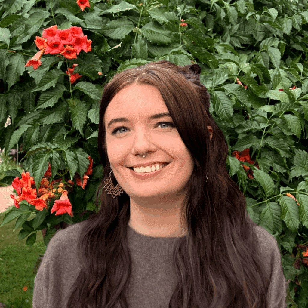 Woman with long dark hair smiling in a lush garden with red flowers and green leaves.