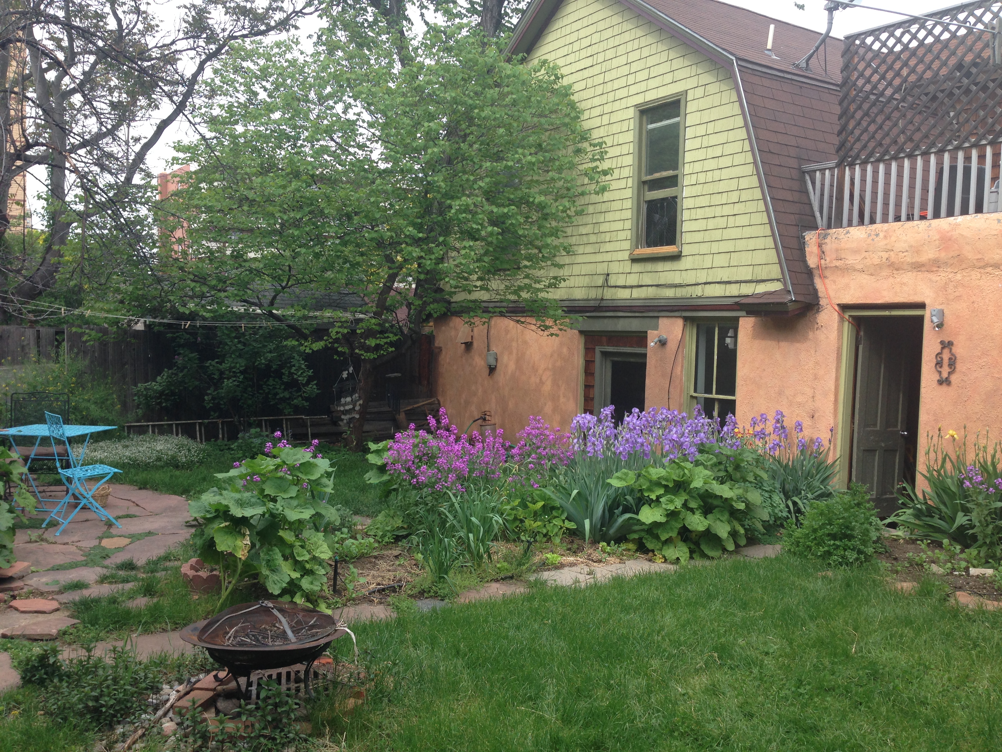 Lush garden with flowers, trees, and a house in the background.