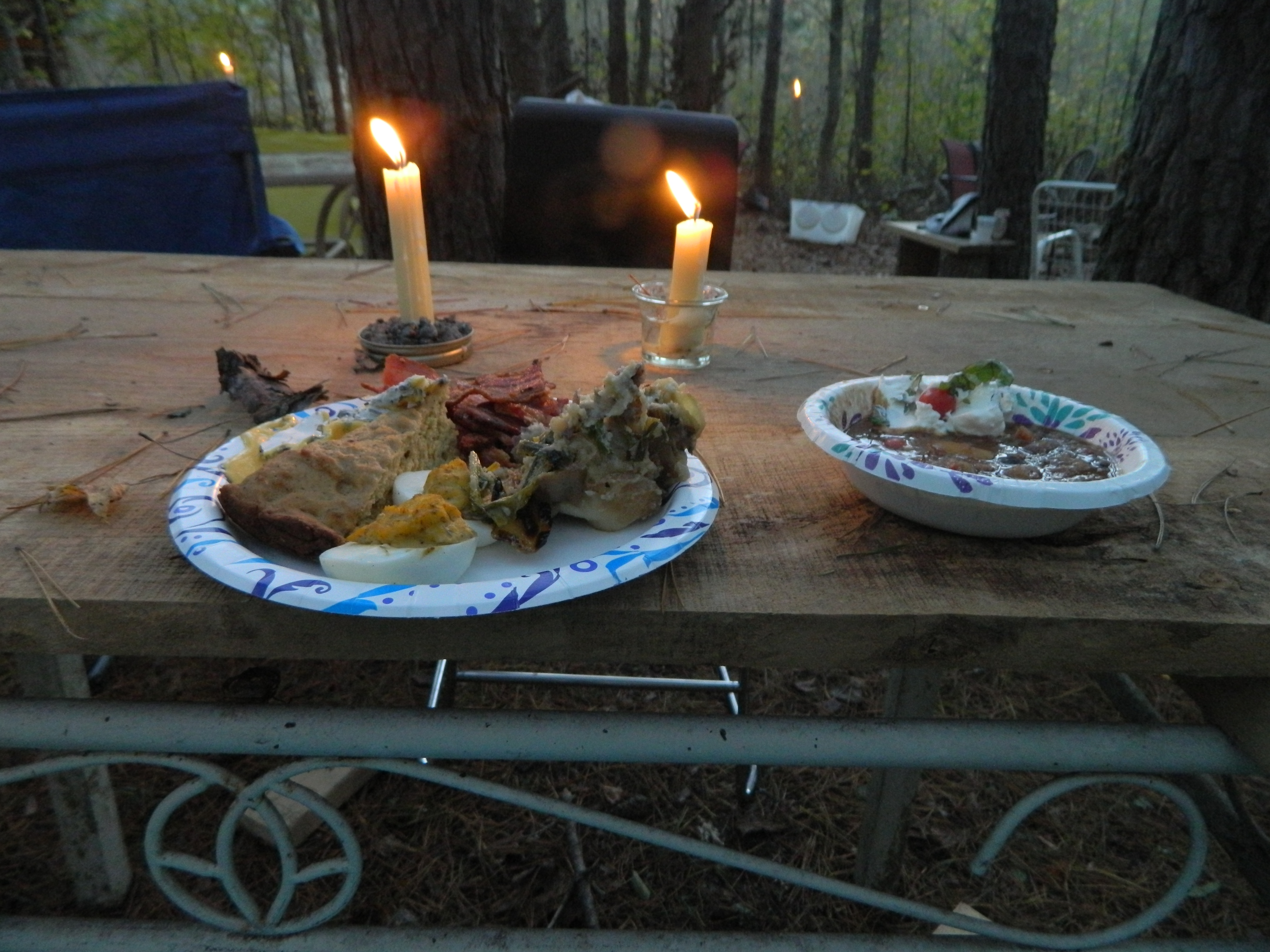 Outdoor dinner setup with candles and food on a wooden table in a forest setting.