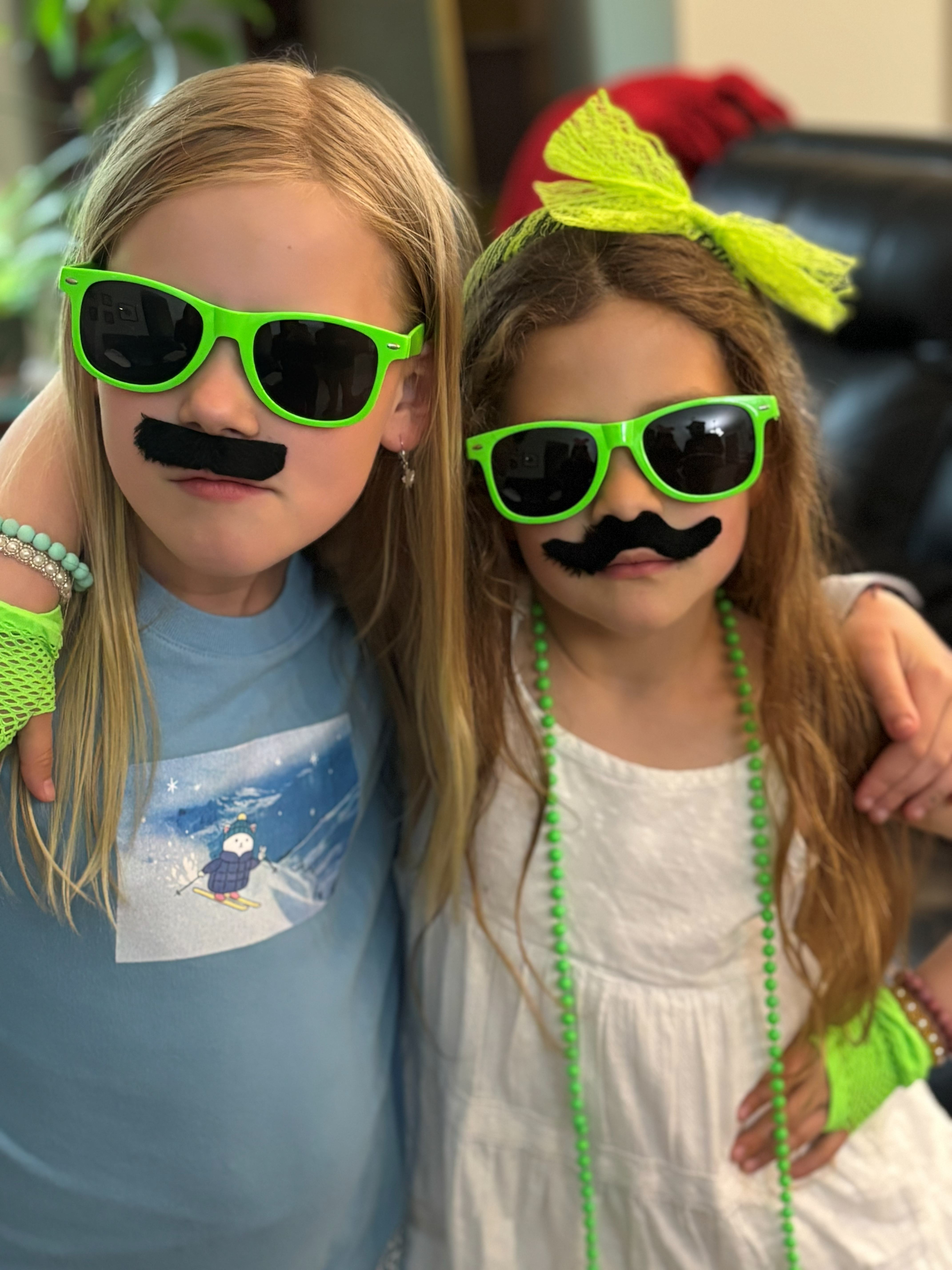 Two kids wearing green sunglasses, fake mustaches, and colorful accessories for a community event.