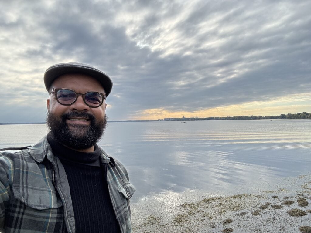 Smiling man with glasses and cap by calm lake during cloudy weather.