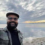 Smiling man with glasses and cap by calm lake during cloudy weather.