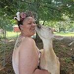 Woman smiling with a baby goat under a tree in a green outdoor setting.