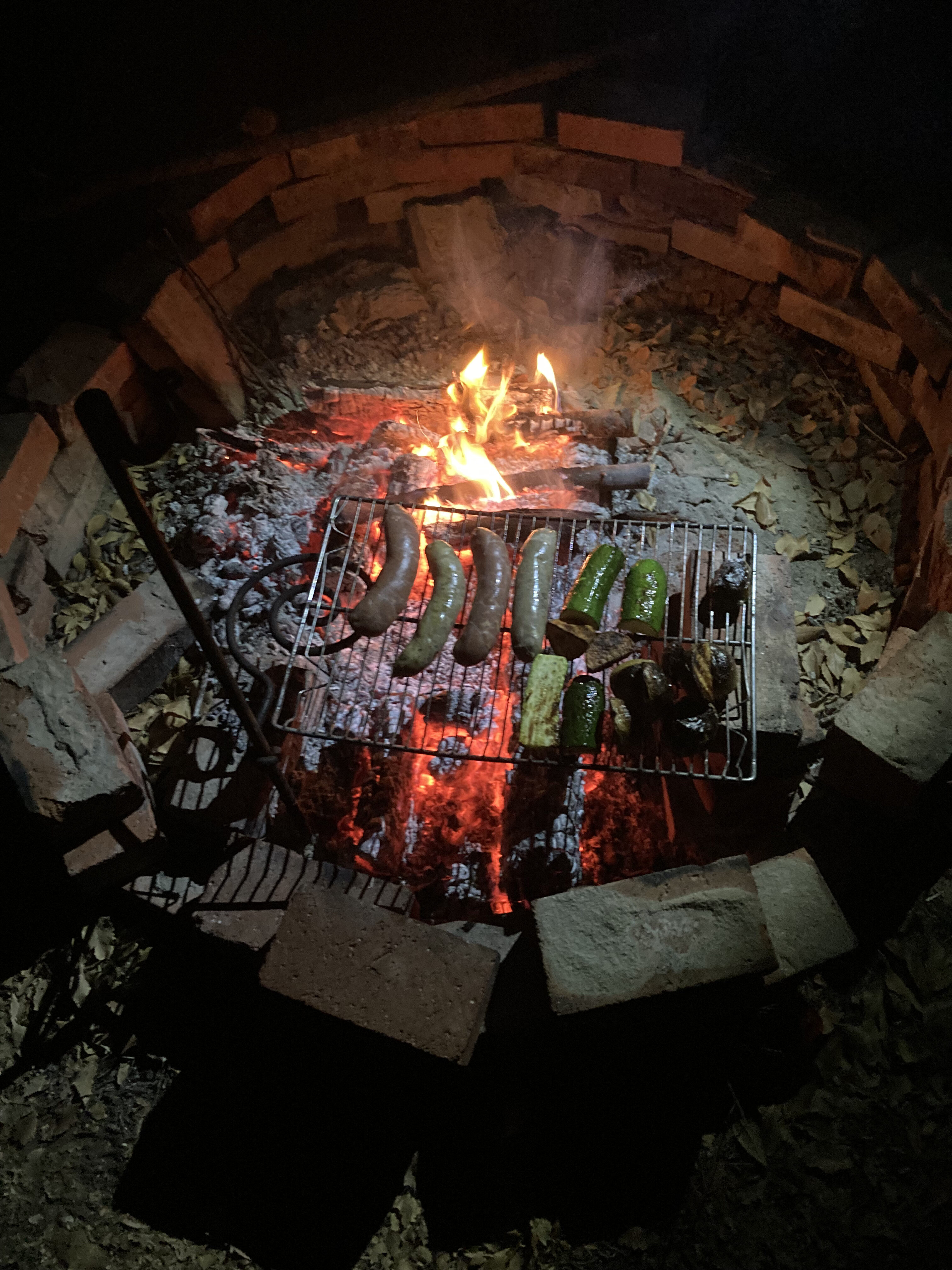 Fire pit with sausages and corn grilling over open flame at night.