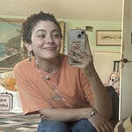 Young woman with short hair smiling while taking a selfie in a cozy, decorated room.