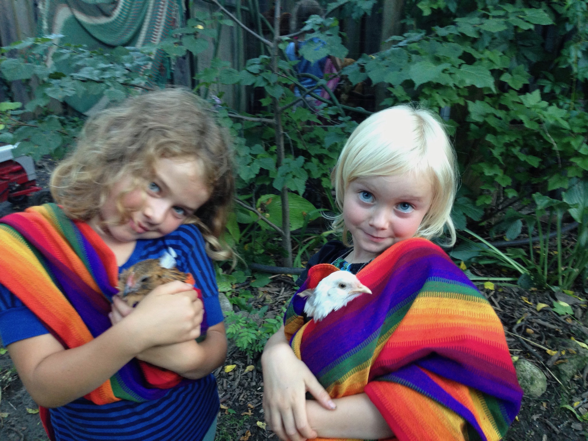 Two children holding guinea pigs wrapped in rainbow-colored fabric outdoors.