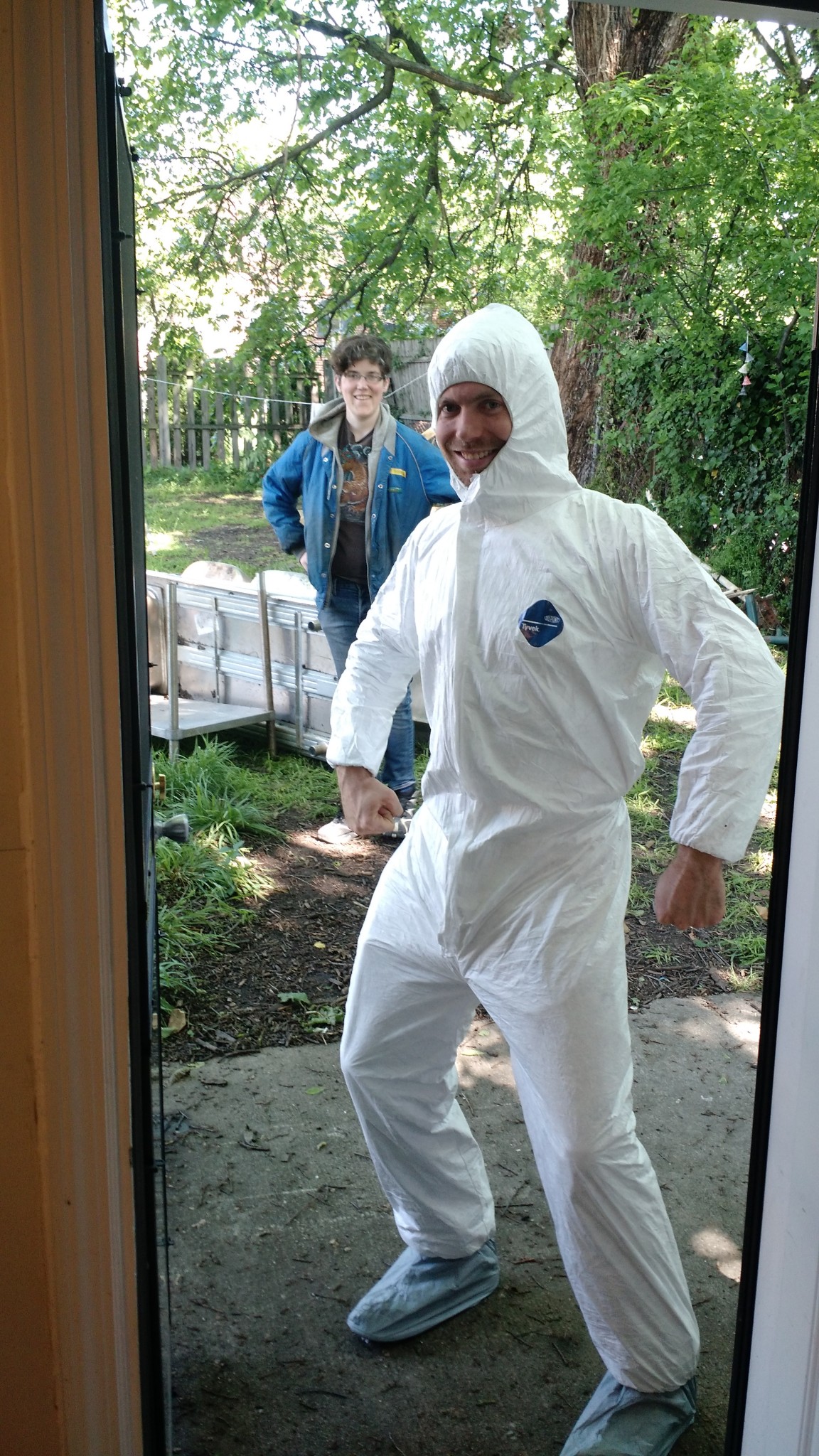 Person in white protective suit standing outside near a door with a person in the background.