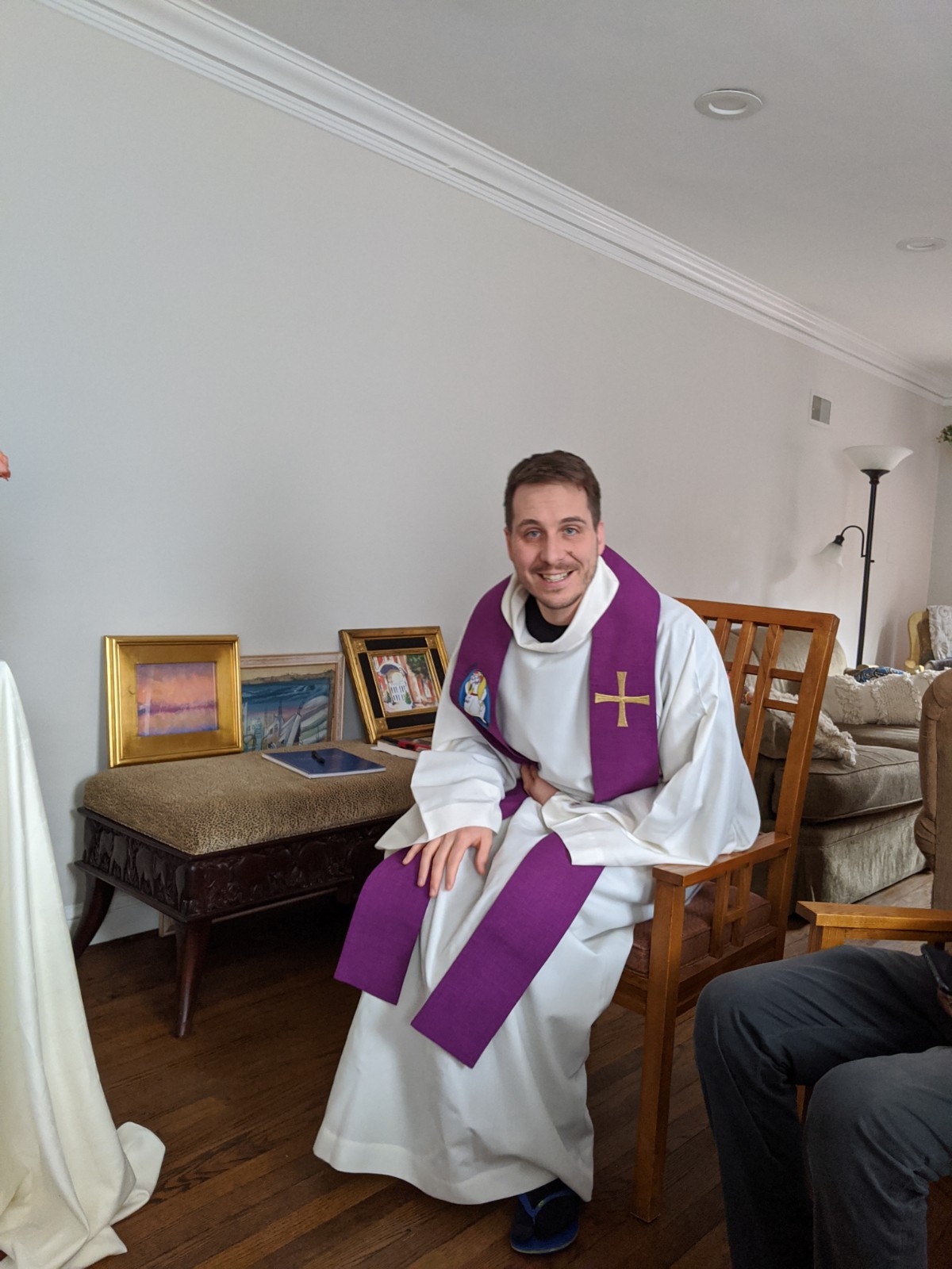 Clergy member in white robe with purple stole smiling indoors.