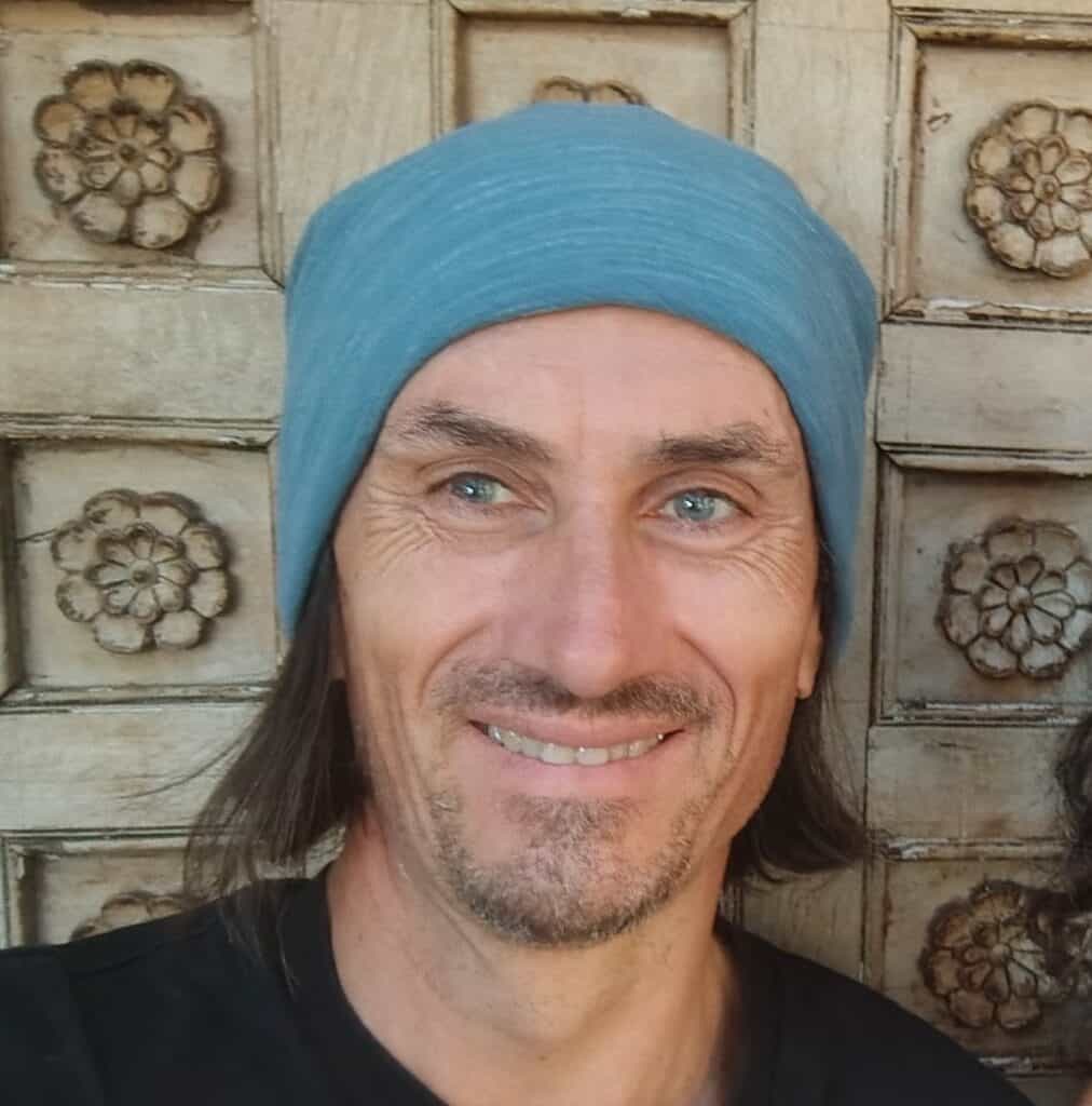 Smiling man wearing a blue headscarf in front of a carved wooden background.