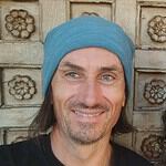 Smiling man wearing a blue headscarf in front of a carved wooden background.