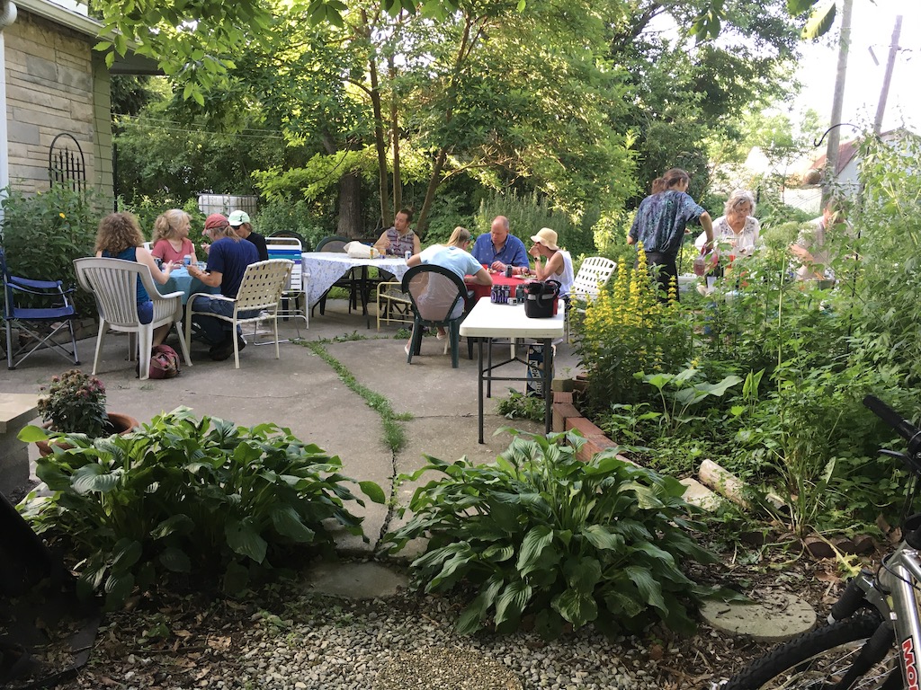 People gathered around tables in a lush garden setting for a community event.