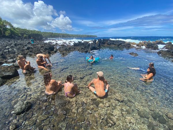 People enjoying a natural tide pool at the Hawaii coast with rocky surroundings and ocean view.