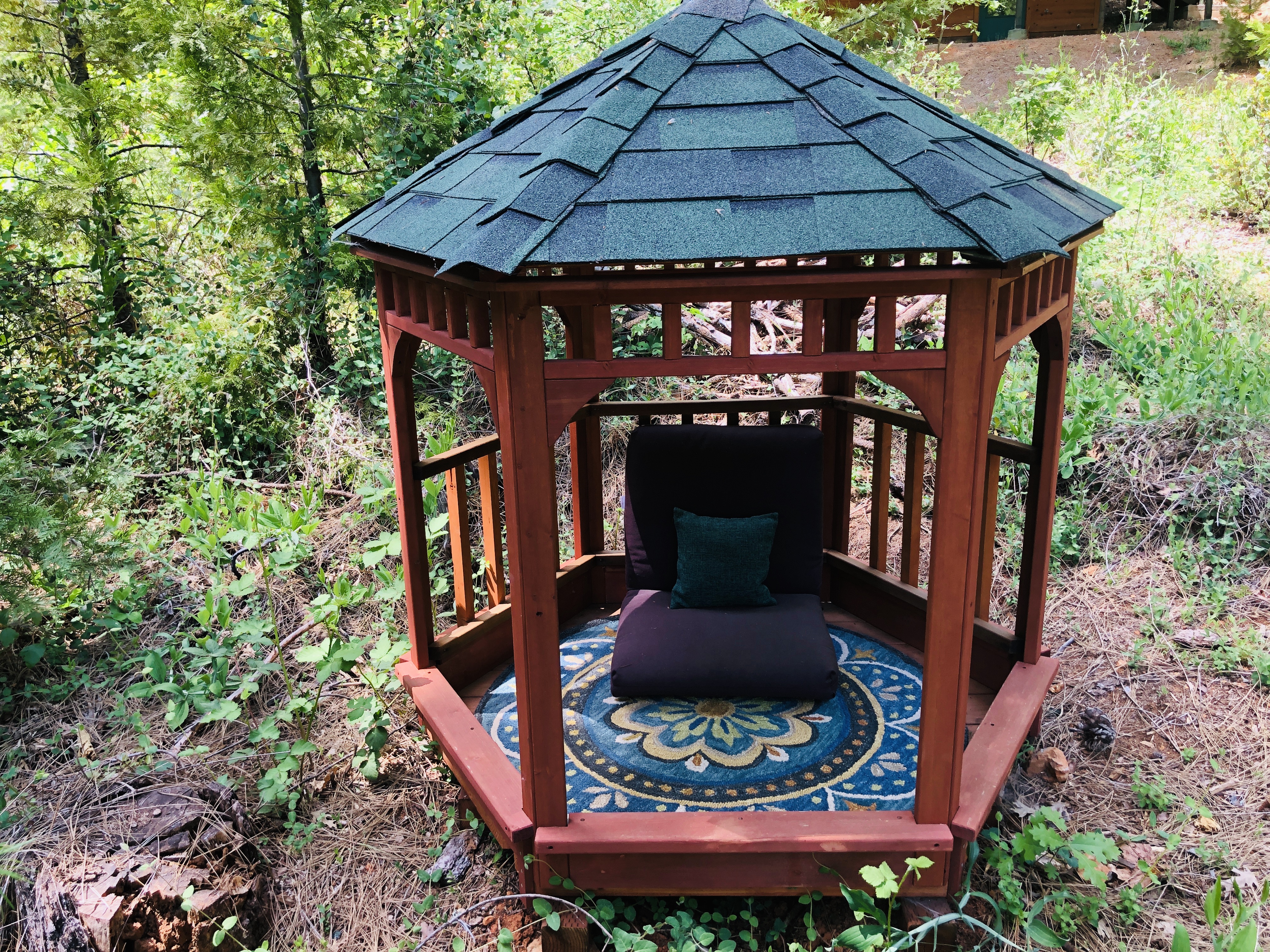 Cozy wooden gazebo in a garden with cushions and a decorative rug.