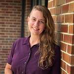 Smiling woman with long wavy hair in purple shirt leaning against brick wall.