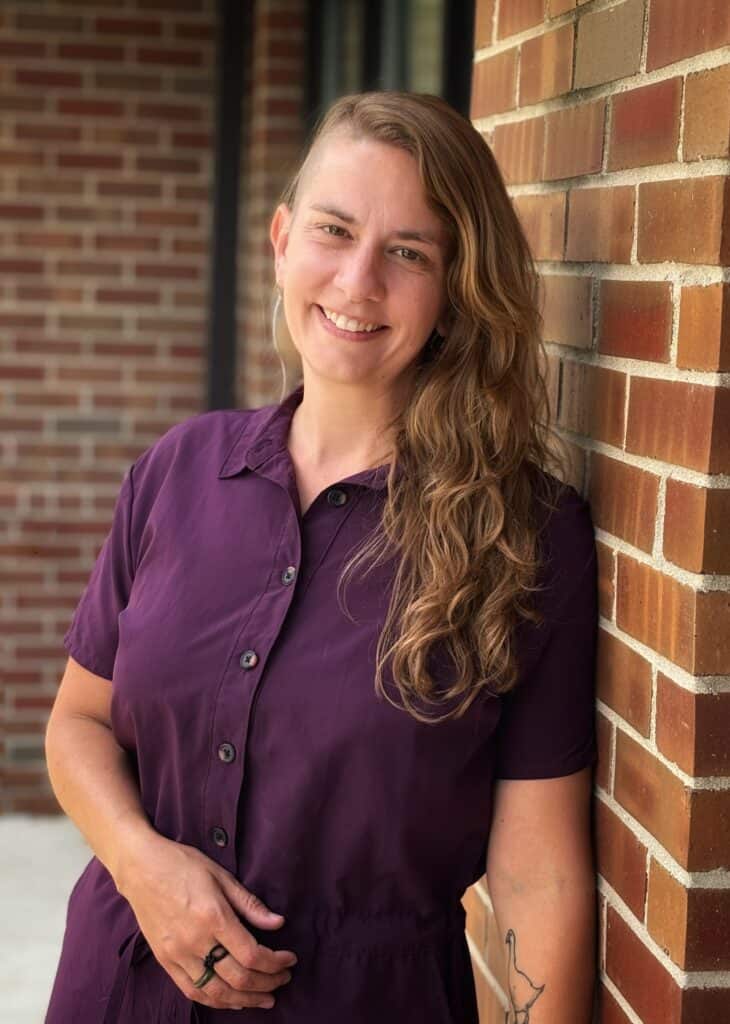 Smiling woman with long wavy hair in purple shirt leaning against brick wall.