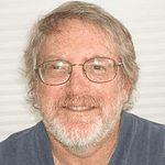 Smiling man with glasses and beard, casual attire, indoor setting.
